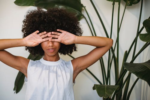 African American female with Afro hairstyle standing near green plant and hiding eyes behind hands