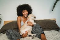 一位快乐的非裔美国女主人抱着白色小狗，坐在舒适的床上，在家度过闲暇时光