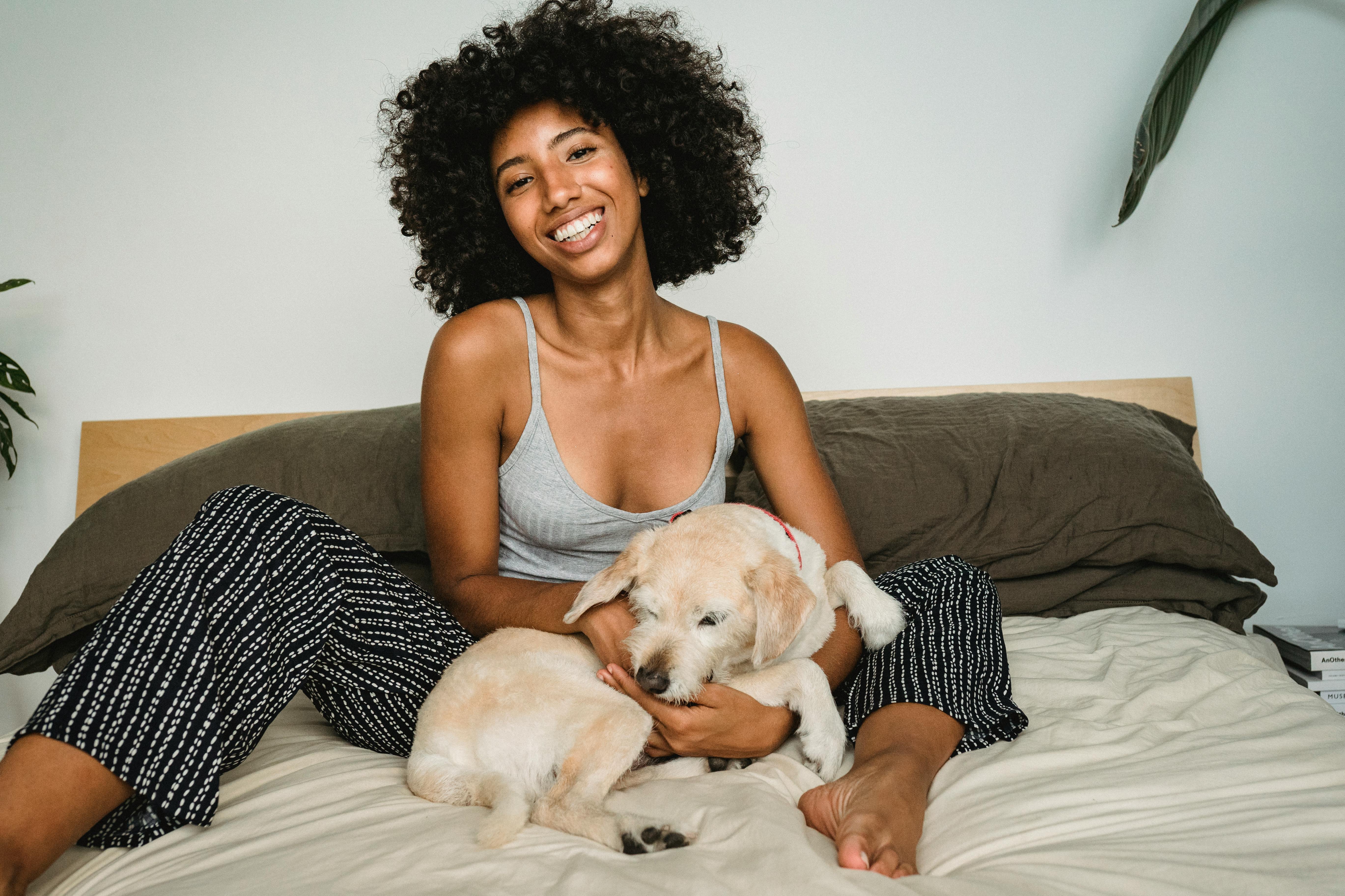 Smiling African American female in casual clothes chilling with small dog in cozy bedroom at home.