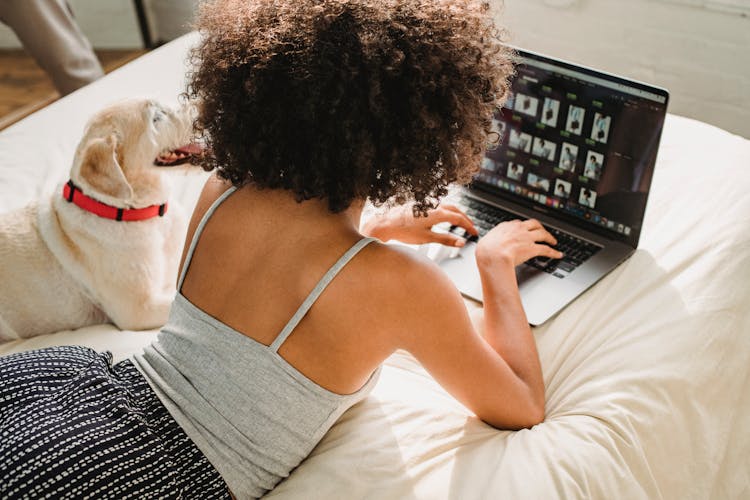 Black Woman Browsing Laptop With Dog On Bed