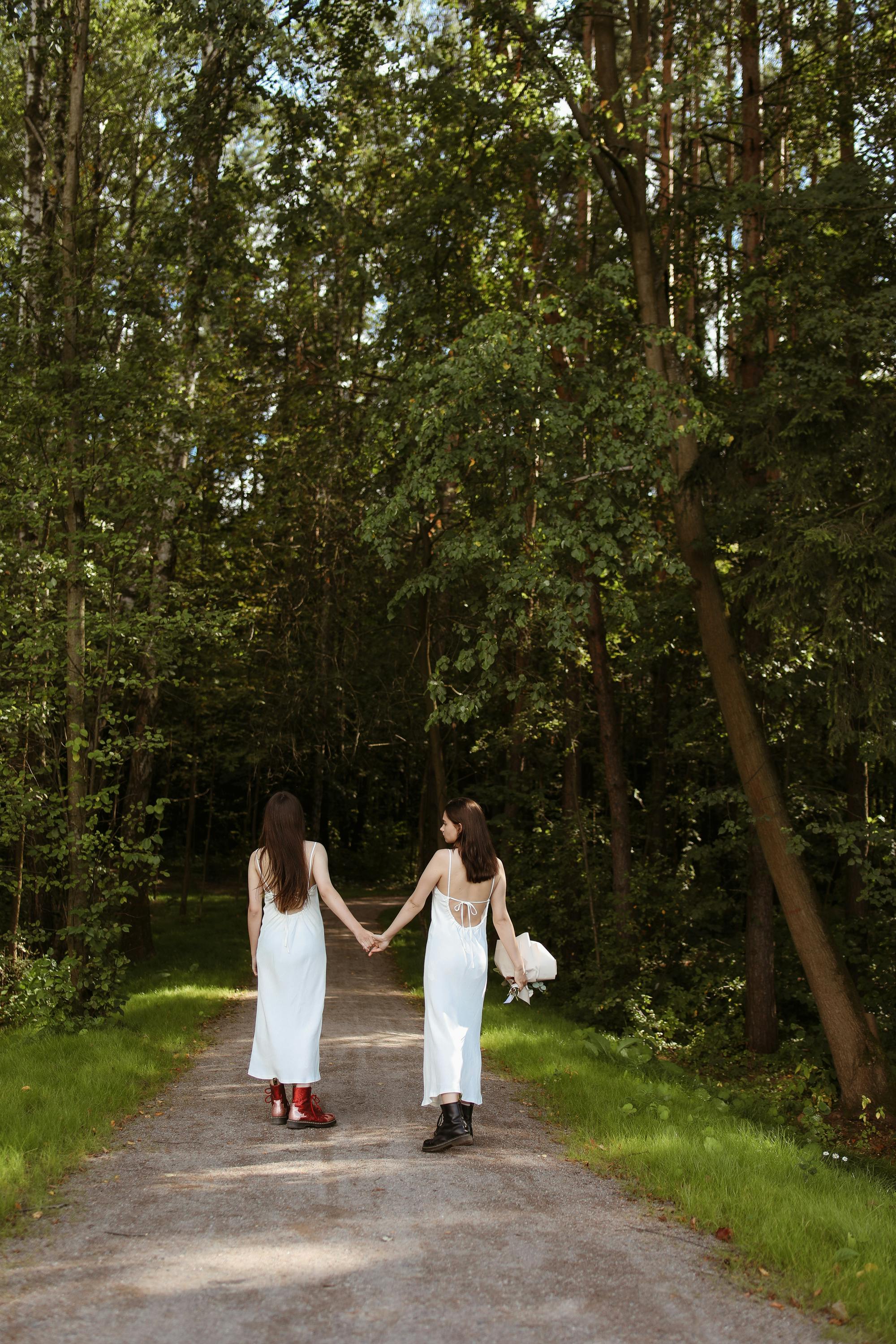 Two women in white dresses holding hands on a forest path, symbolizing love.
