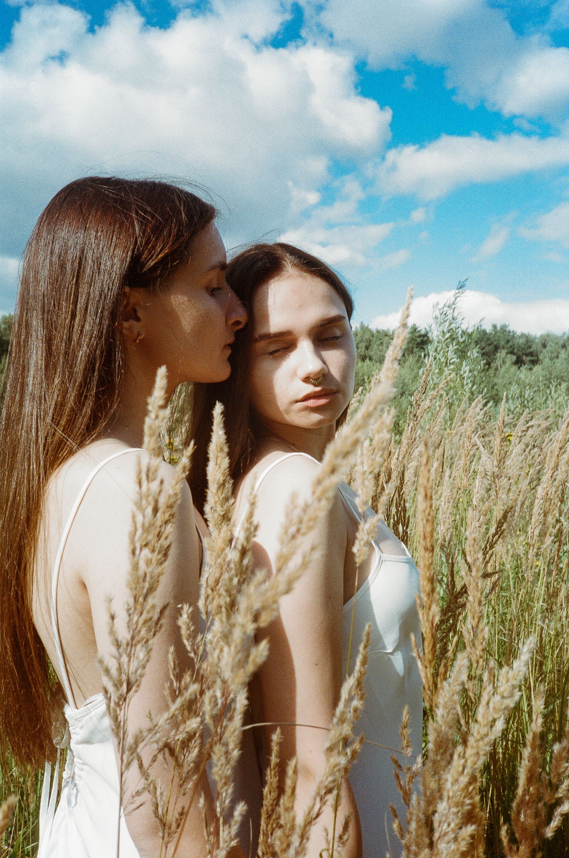 Two women in a sunny field, embracing affectionately, in white dresses.