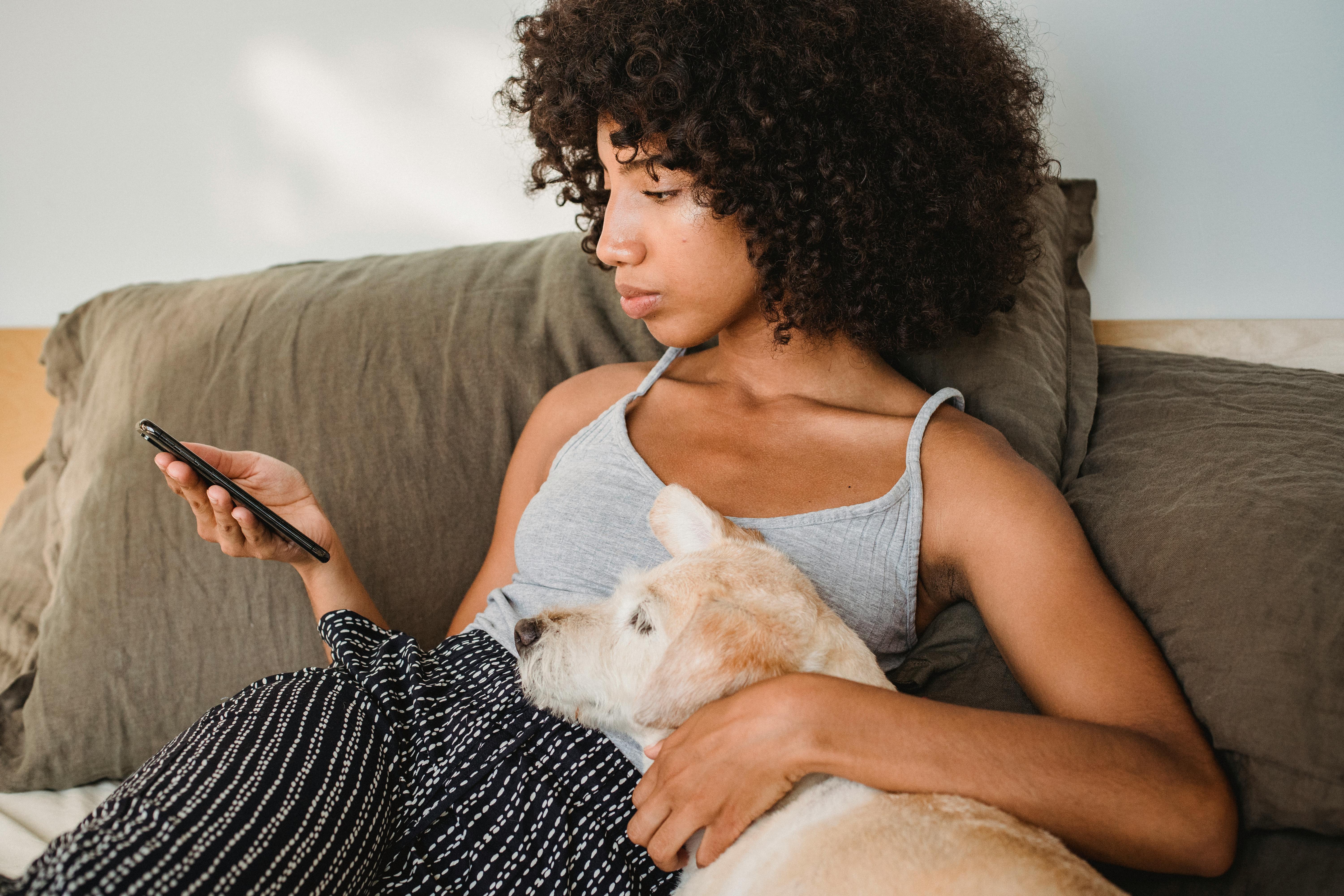 crop black woman watching smartphone while embracing dog on bed