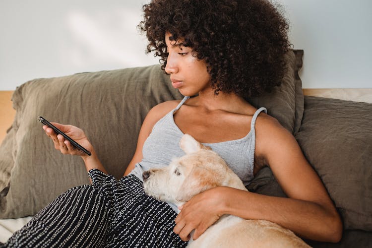 Crop Black Woman Watching Smartphone While Embracing Dog On Bed