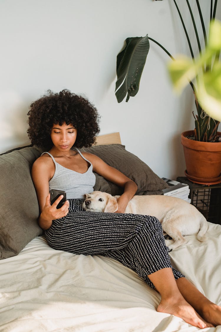 Ethnic Woman Watching Smartphone With Dog On Bed