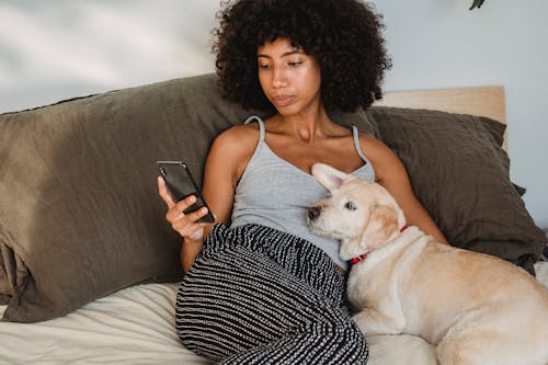 Crop black woman with smartphone lying on bed with dog
