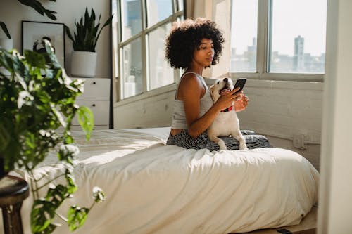 Side view of young ethnic female watching cellphone while sitting on bed with dog at home