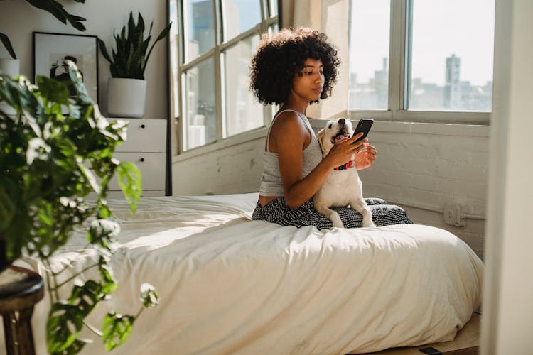 Black Woman With Smartphone And Dog On Bed In House