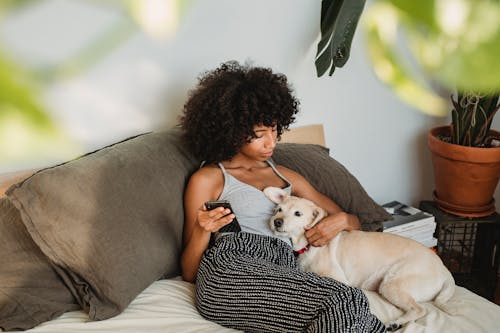 Ethnic woman with dog and smartphone resting on bed