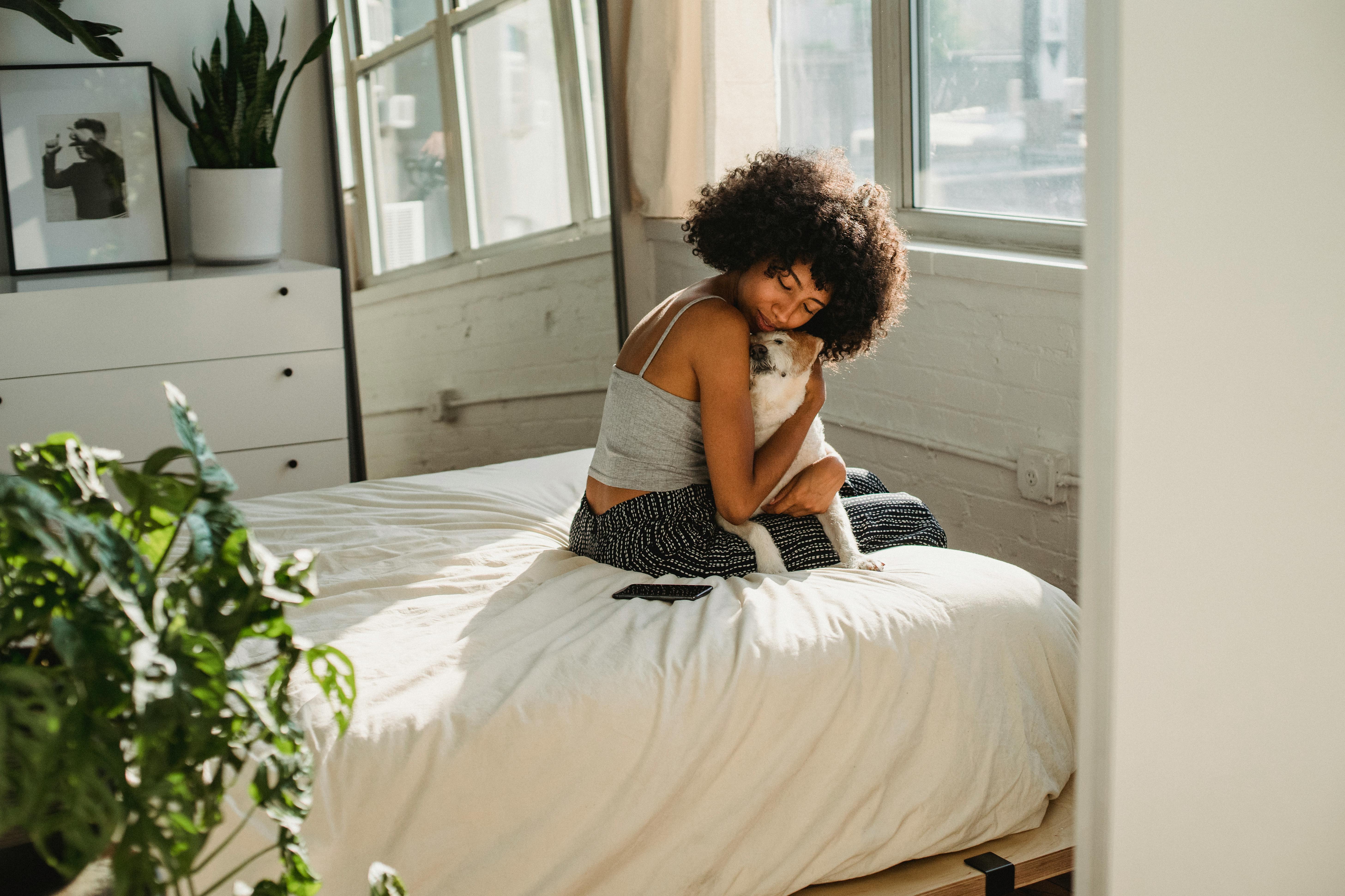 Side view of ethnic female with closed eyes cuddling purebred dog on soft bed in house