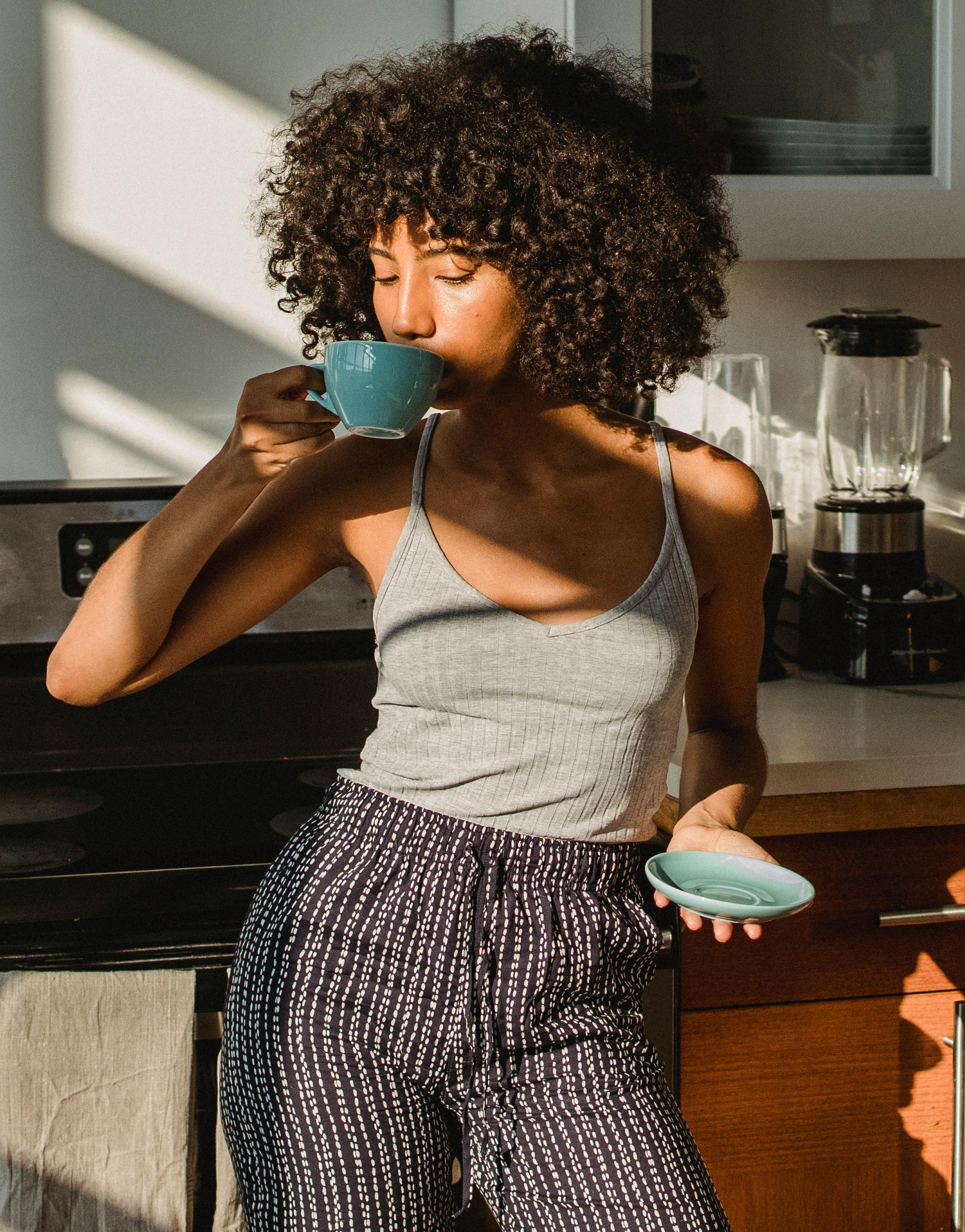 Young ethnic female with Afro hairstyle drinking hot beverage from cup in house in sunlight