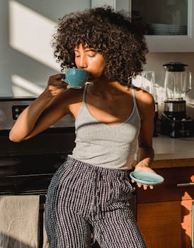 Young African American woman in casual attire savoring a hot drink in a sunlit kitchen.