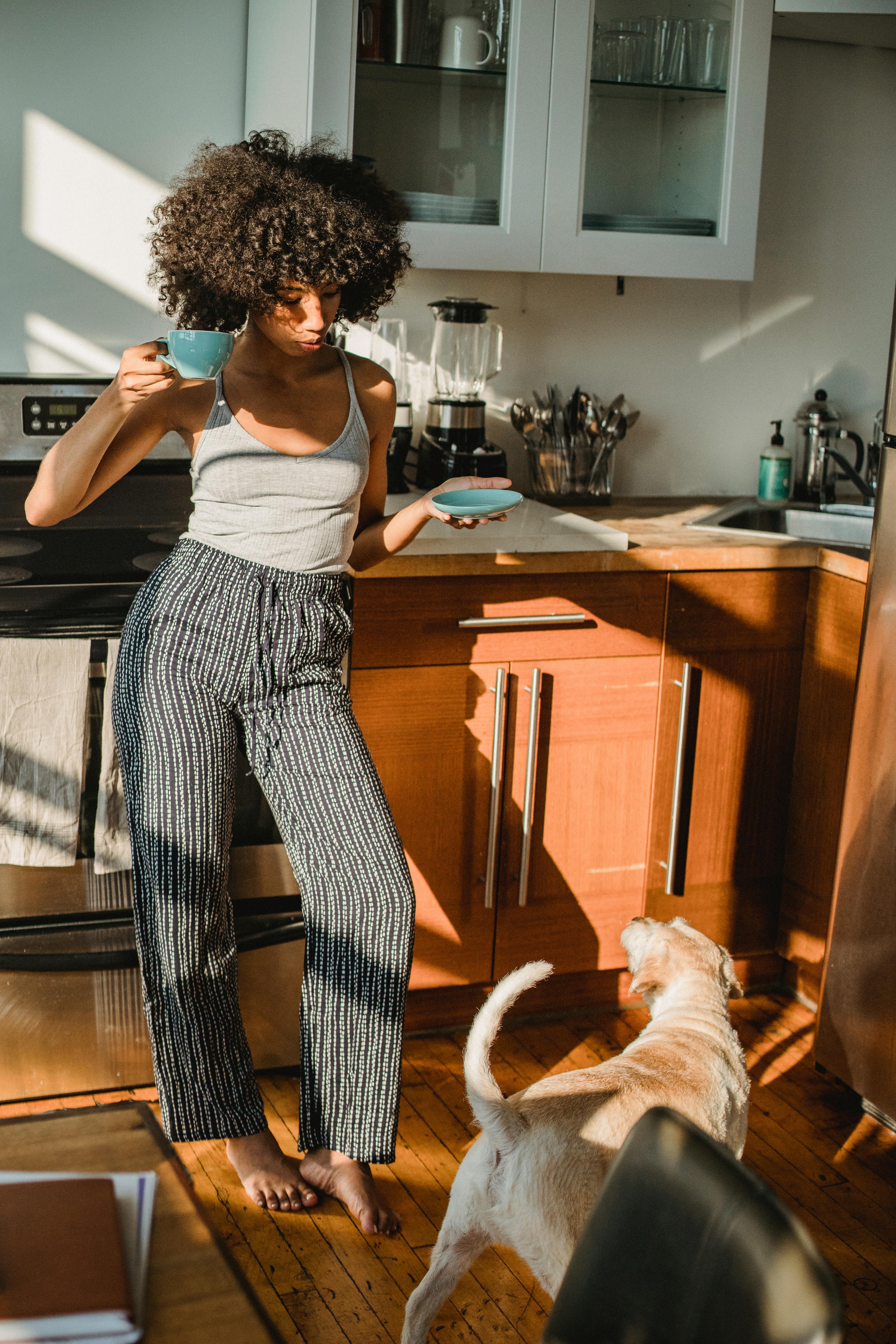 一位穿着休闲装的年轻非裔美国女性，手里拿着一杯热饮，在阳光下站在家里的狗旁边