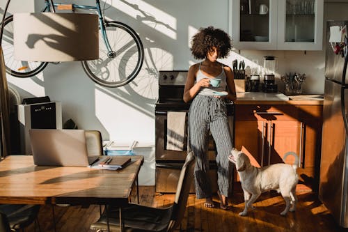 Young barefoot African American female with cup of hot drink standing on floor with dog at home