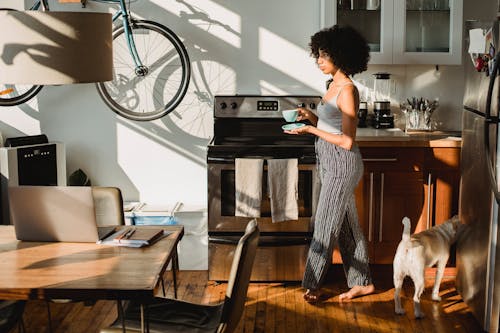 Ethnic woman with hot drink walking on floor near dog