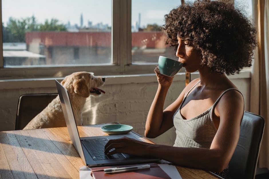 Side view of concentrated young African American lady sitting at table in home while working remotely on computer and drinking coffee near big dog