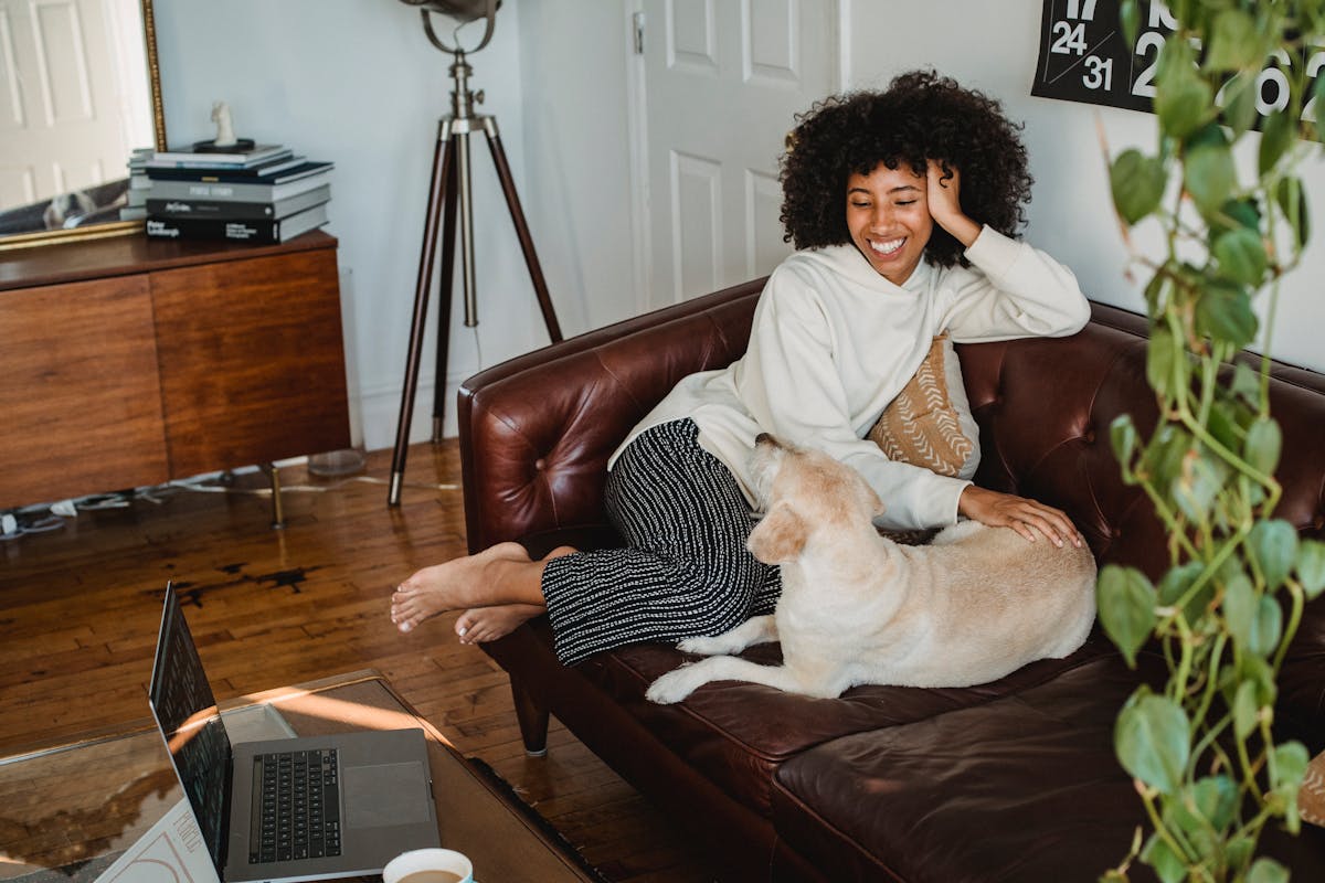 Happy young African American woman sitting on couch and resting with adorable dog with computer and cup of coffee on table in living room
