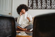 Young African American lady in casual clothes resting on sofa with dog and speaking on smartphone with notepad in legs in living room