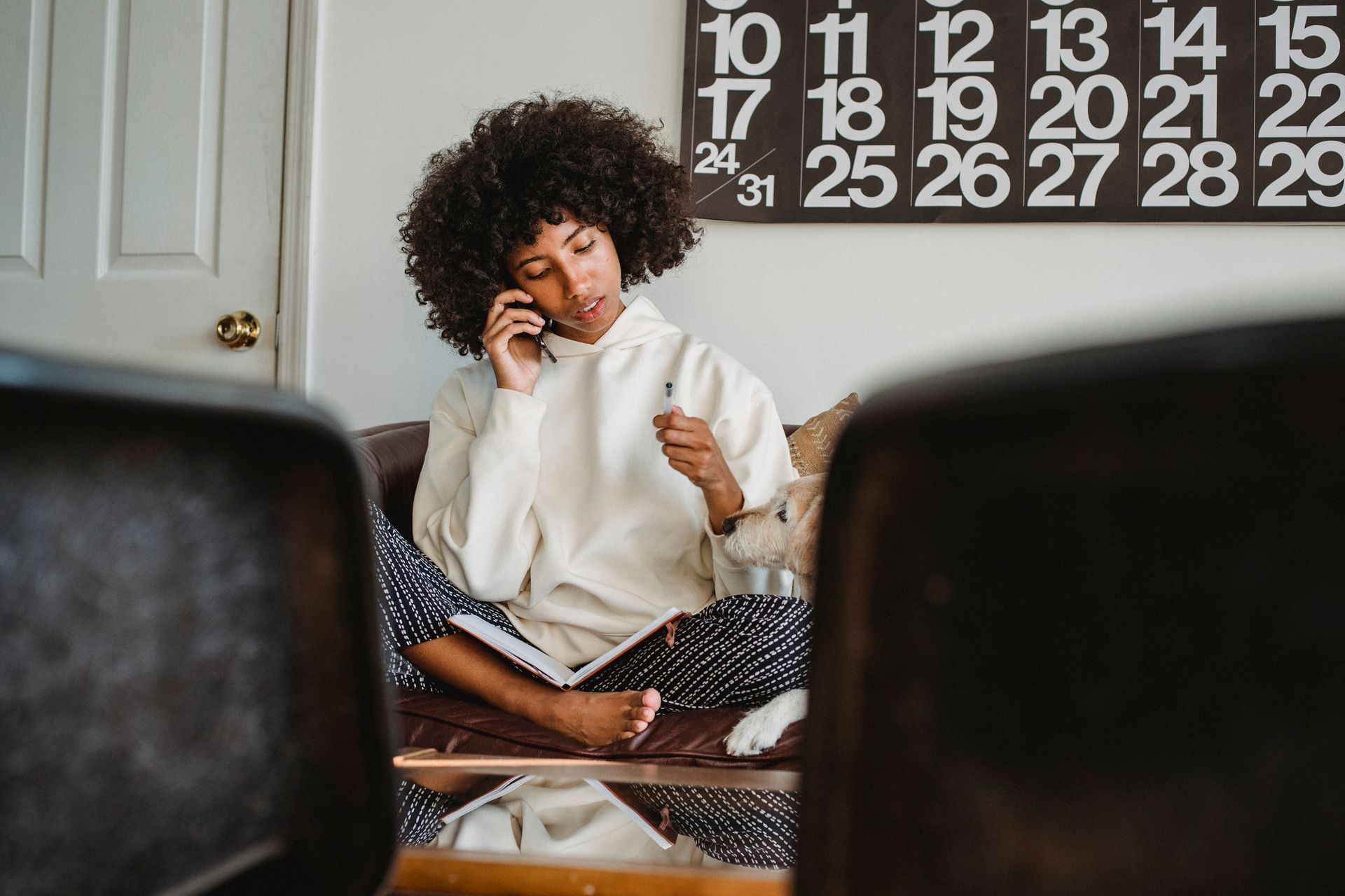 Young African American lady in casual clothes resting on sofa with dog and speaking on smartphone with notepad in legs in living room