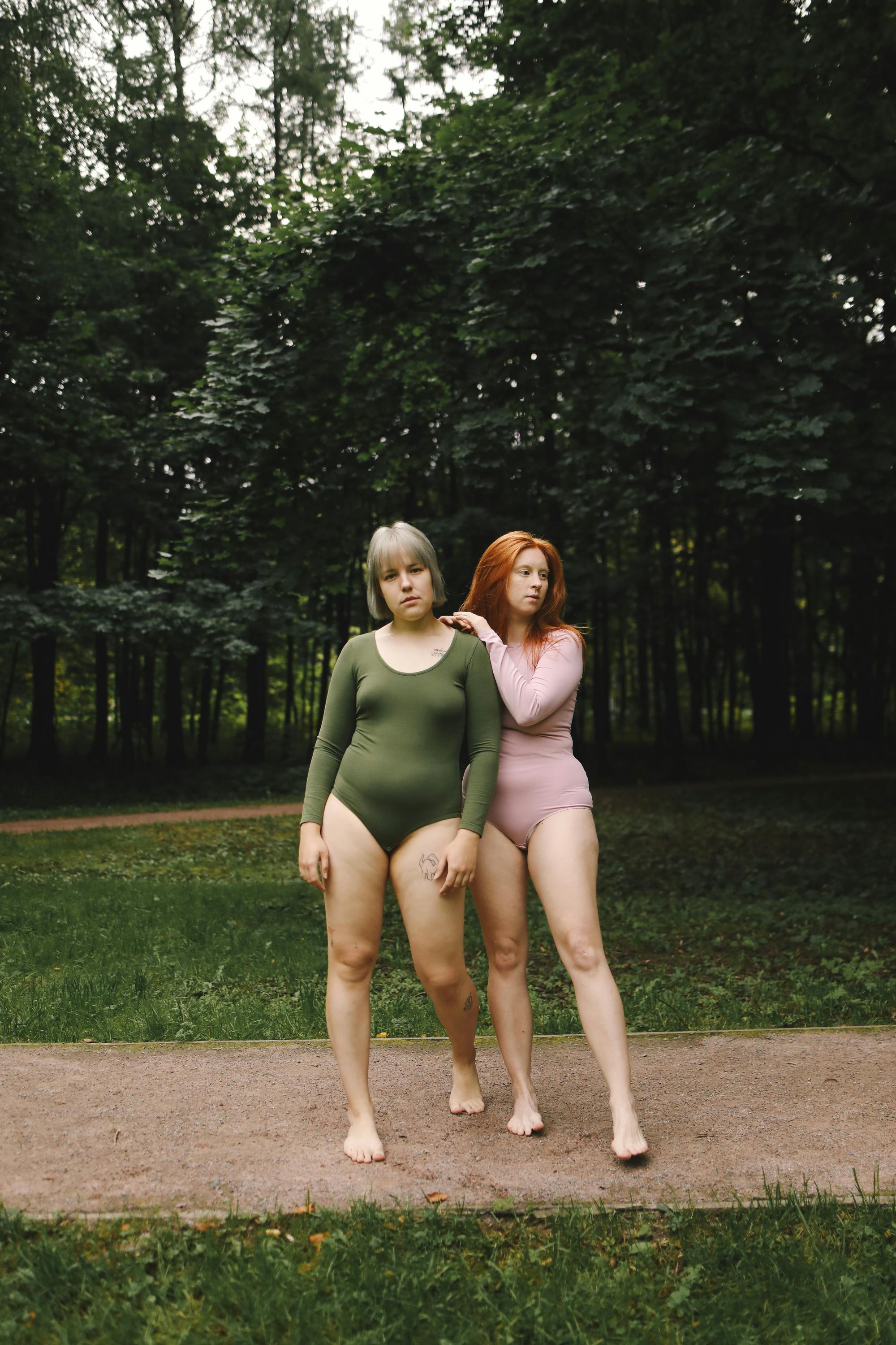 Two women in bodysuits standing barefoot in a lush green park.