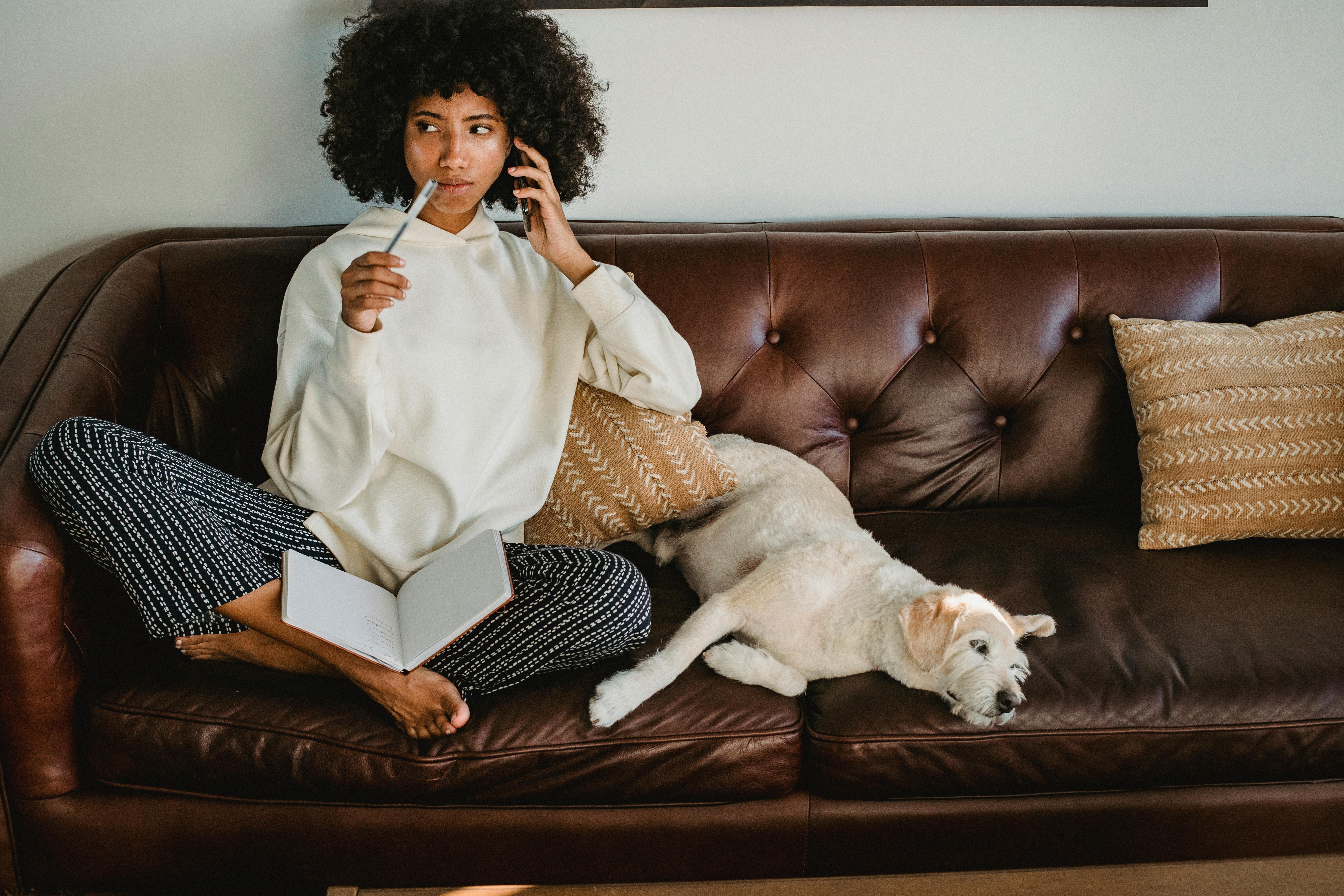 thoughtful black woman with notebook and dog on leather sofa