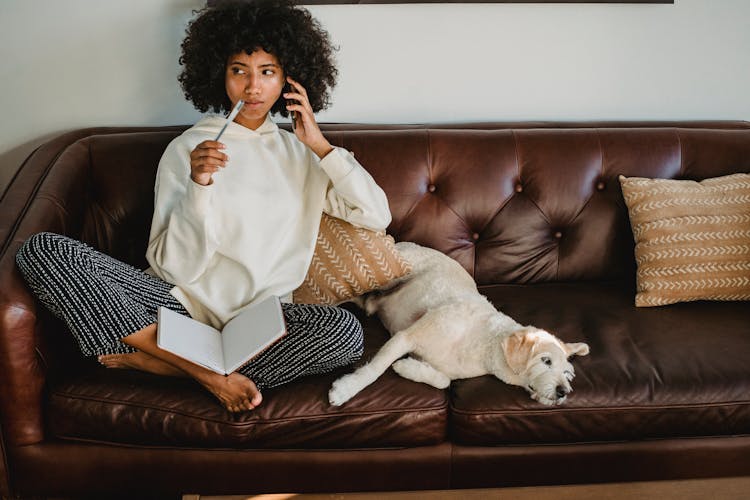 Thoughtful Black Woman With Notebook And Dog On Leather Sofa