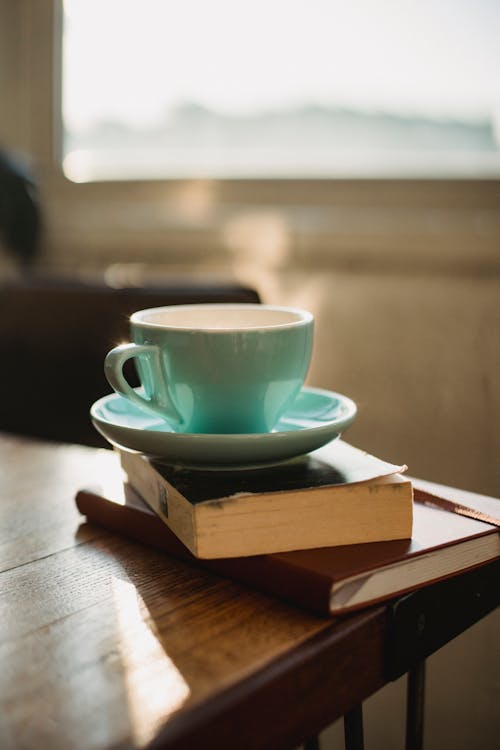 Cup of fresh aromatic coffee and books on wooden table against window at home