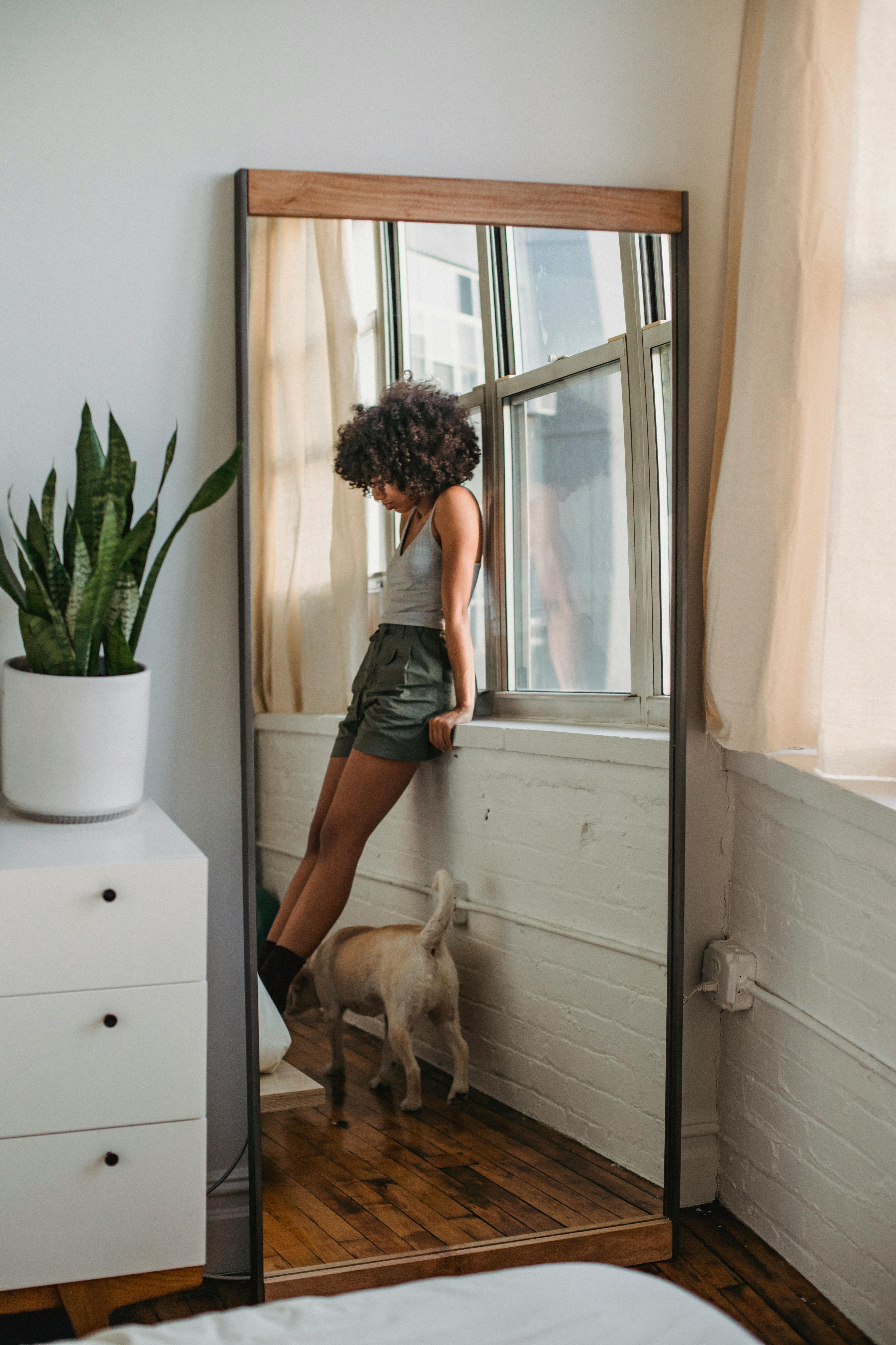 一位留着非洲发型的无脸黑人女性，身穿休闲服，靠在窗台上，与可爱的小狗共度时光