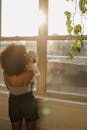 Back view of anonymous black female in domestic clothes embracing cute puppy while standing near window and enjoying sunny morning