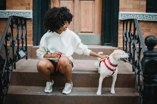 Full body of cheerful African American female in trendy outfit stroking adorable puppy with leash while resting on steps