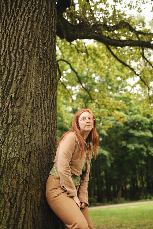 A Woman in Brown Jacket and Pants Leaning on Tree Trunk