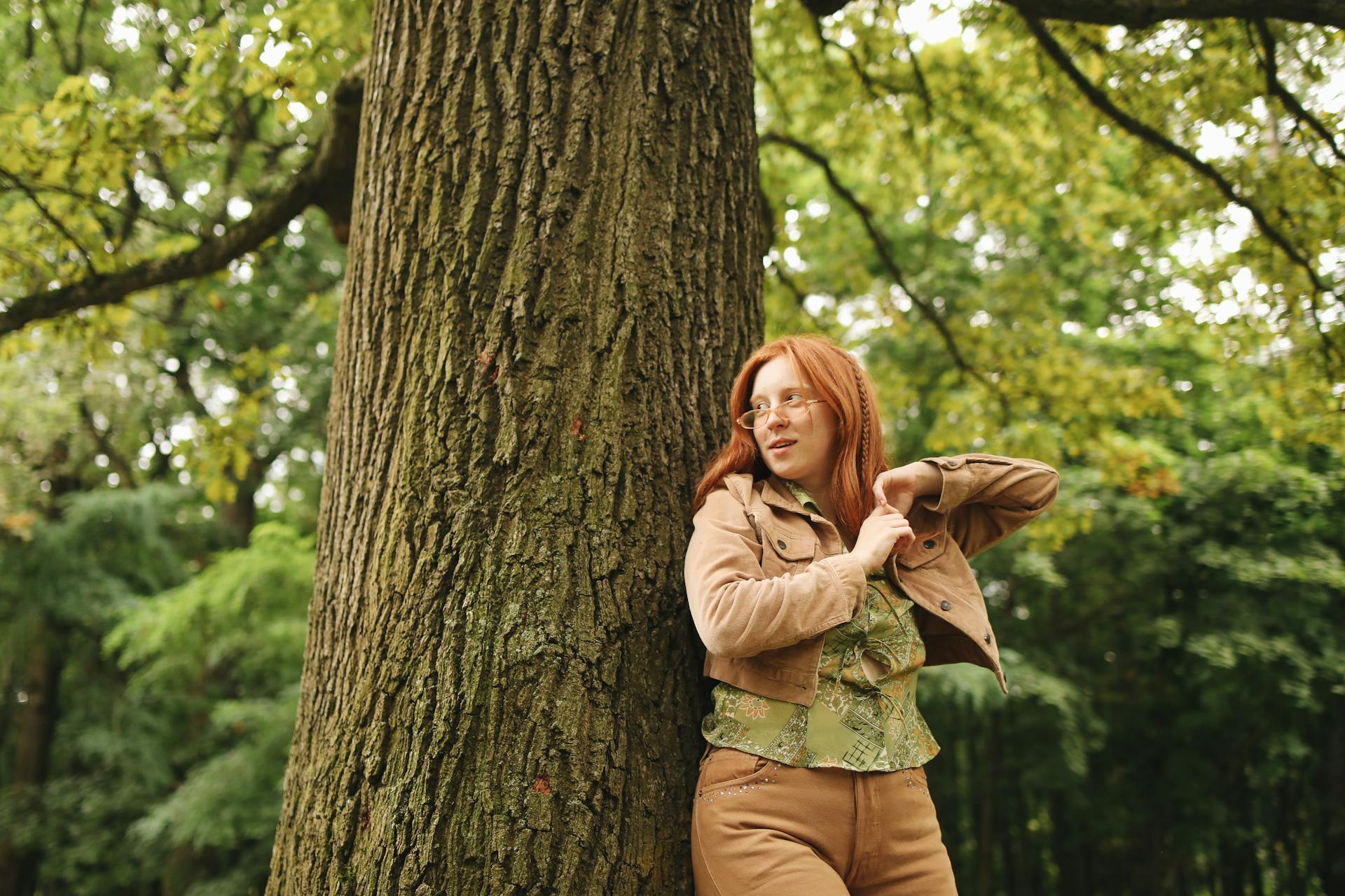 Smiling woman with red hair and glasses leaning on a large tree in a lush green forest.