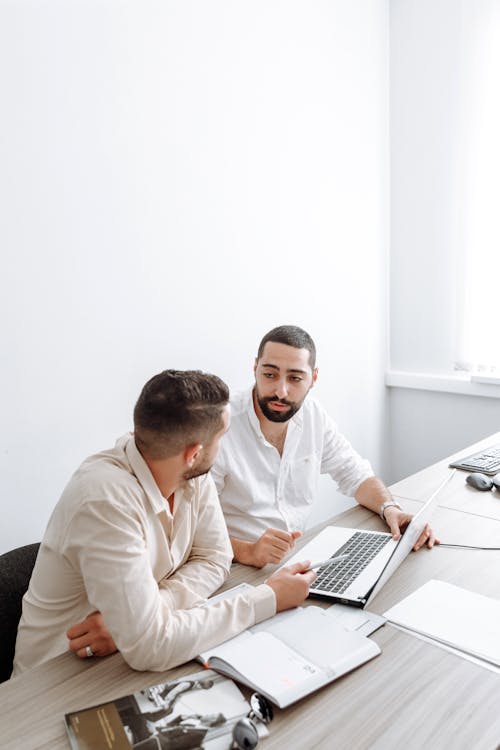 Two Men in White Long Sleeves Talking Beside a Laptop