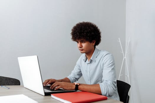 A young man with curly hair intently working on a laptop in a modern office setting.