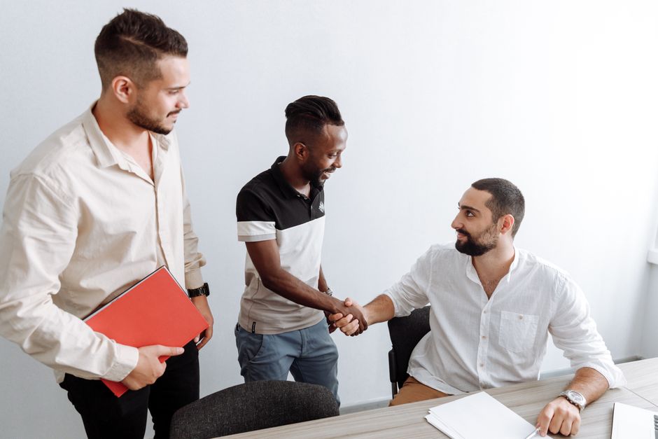 Three men in an office setting, shaking hands, representing teamwork and diversity. Three men in an office setting, shaking hands, representing teamwork and diversity.