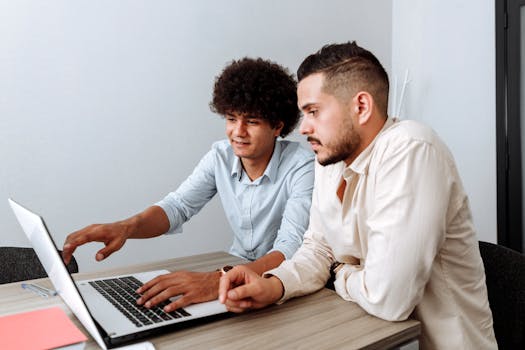 Two young men working together on a laptop in a modern office setting.