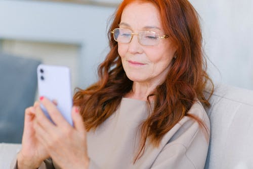 Free Aged female with red hair in eyeglasses looking at screen of smartphone and making online video call on blurred background Stock Photo