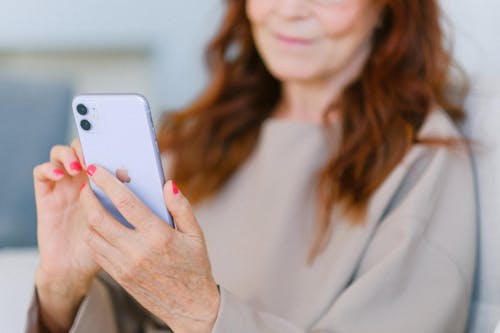 Crop anonymous aged female with red hair looking at screen of modern mobile phone and touching screen in soft focus