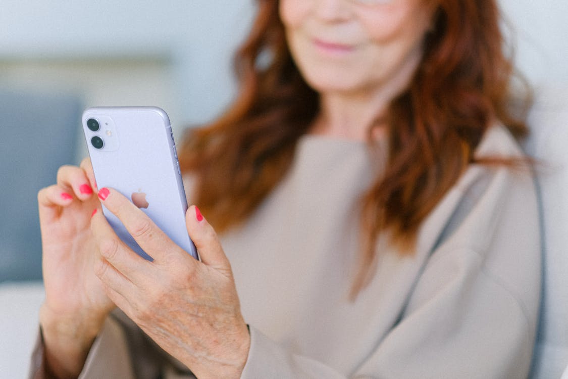 Crop anonymous aged female with red hair looking at screen of modern mobile phone and touching screen in soft focus
