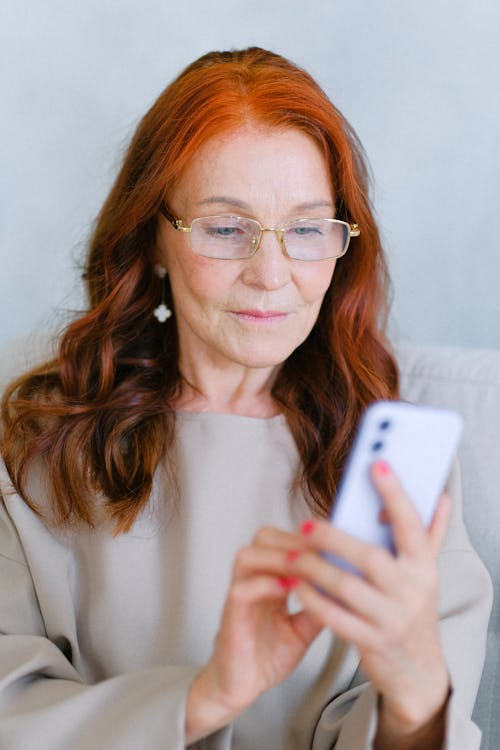 Elderly redhead woman in eyeglasses texting on smartphone