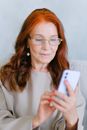 Elderly redhead woman in eyeglasses texting on smartphone
