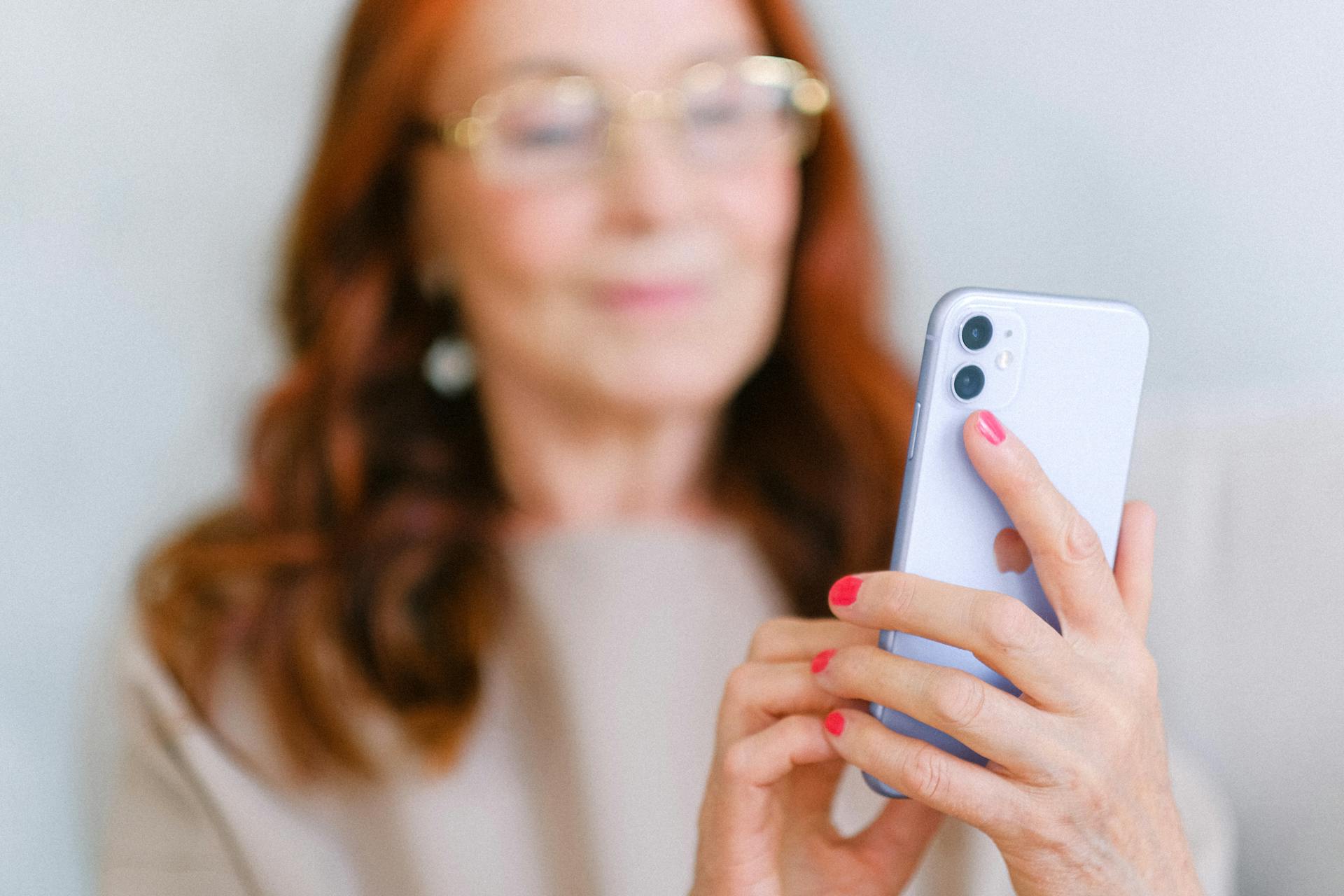 Crop aged redhead female in eyeglasses using contemporary mobile phone while surfing internet in soft focus