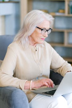 Elderly woman with glasses typing on a laptop in a cozy home setting, embodying remote work lifestyle.