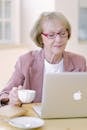 Aged female in eyeglasses with cup checking important documents on netbook on blurred background