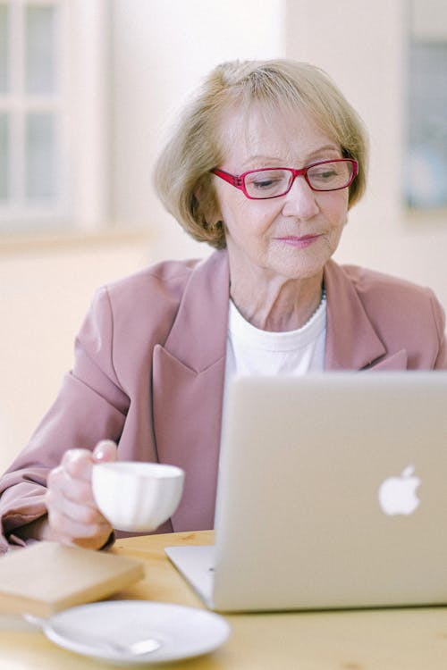 Free Aged female in eyeglasses with cup checking important documents on netbook on blurred background Stock Photo