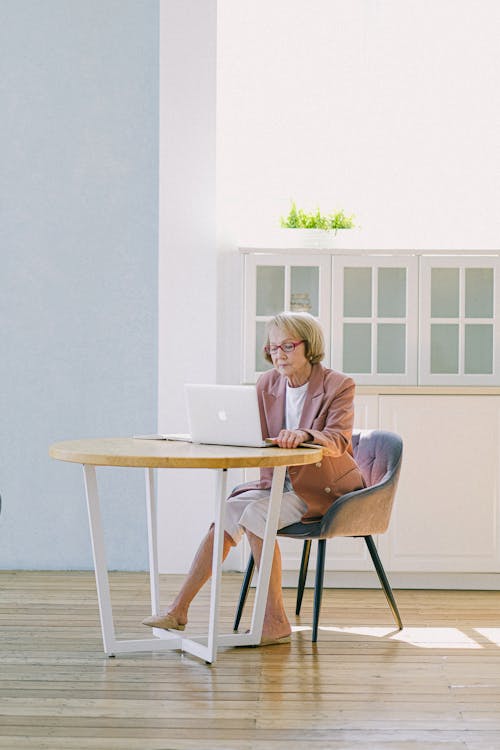 Full body of elderly female in eyeglasses improving important report while using netbook at table in modern room