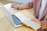 From above of crop anonymous elderly female in elegant jacket with pen in hand using netbook and reading book