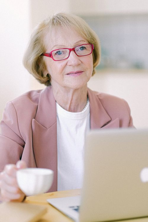Free Elderly female in eyeglasses with mug and netbook looking at camera while working at table on blurred background Stock Photo
