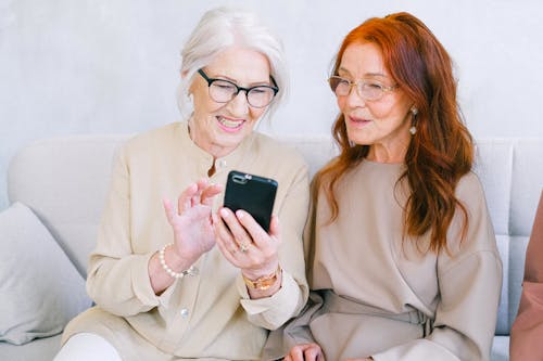Free Happy elderly elegant trendy women looking at screen of mobile phone and shopping online while resting on sofa Stock Photo
