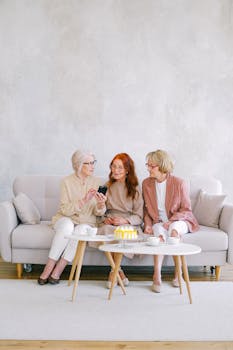 Three senior women enjoying tea and conversation indoors, looking at a smartphone together.